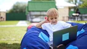 Lovely blond kid using gadgets outdoors. Nice little boy sitting in bean bag chair watching videos on laptop smiling. Close up. Blurred backdrop.