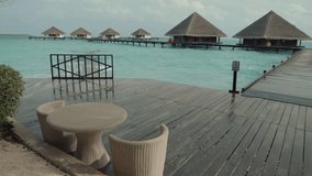 View of beach and cottages with triangle roofs of a luxury Maldives resort with selective focus on a wicker cafe table with two armchairs, the ocean waves crash on a wooden pier in the background
