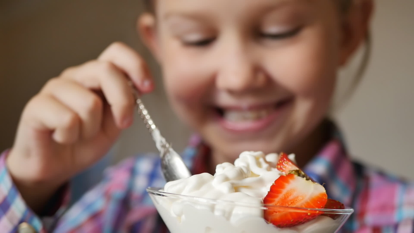 portrait of a cute teenage girl in a plaid shirt eats creamy ice cream with strawberries holding a glass ice cream maker on a leg and a silvery metal spoon in her hands. The child smiles while eating Royalty-Free Stock Footage #1094456593