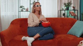 Excited adult girl sitting on sofa eating popcorn and watching interesting tv serial, sport game, film, online social media movie content online at home. Young woman enjoying domestic entertainment