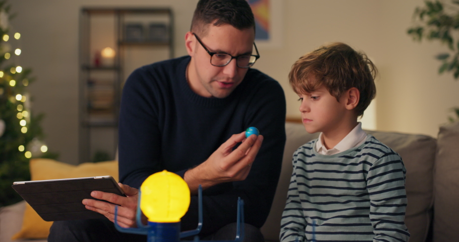 Son is spending time with dad men are sitting on couch playing with a model of planetary system guy is explaining to boy about construction of cosmos using toy as example he is naming planets.