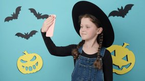 Happy fun little child girl in black hat get video call using mobile cell phone talk greet with hand, isolated on blue color background in studio decorated paper pumpkin and bats. Halloween concept
