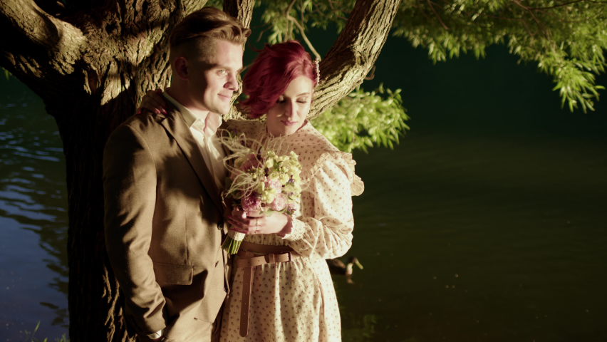beautiful couple of man and woman stand on shore of lake or river and kiss each other. groom in suit and bride in dress at wedding photo shoot in park in nature.
