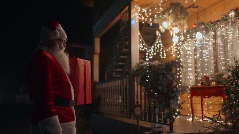 Santa Claus carries the gift in his hands and enters the children's house to leave gifts under the Christmas tree. Decorated Christmas House: stockvideo