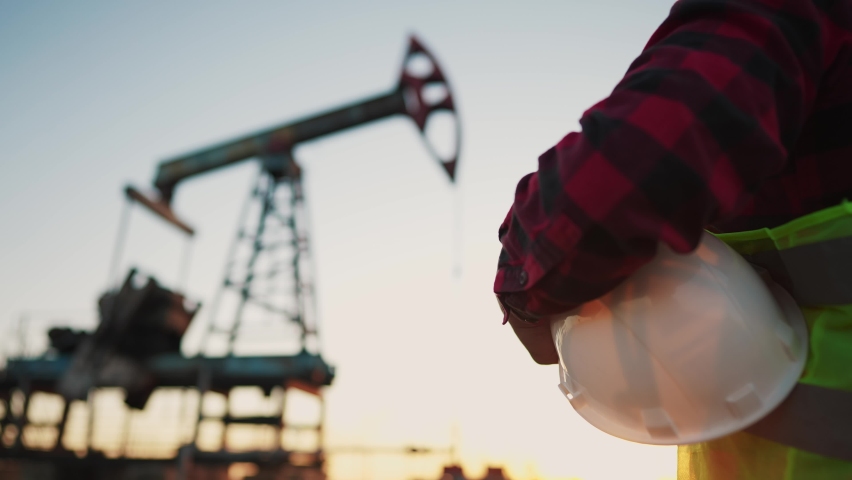 oil production. a worker holding a protective hard hat at sunset in the background an oil pump. oilfield business a extraction concept. oil extraction pump. lifestyle oil pump rig Royalty-Free Stock Footage #1094463659