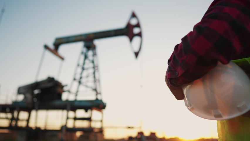 Oil production. a worker holding a protective hard hat at sunset in the background an oil pump. oilfield business a extraction concept. oil extraction pump. lifestyle oil pump rig