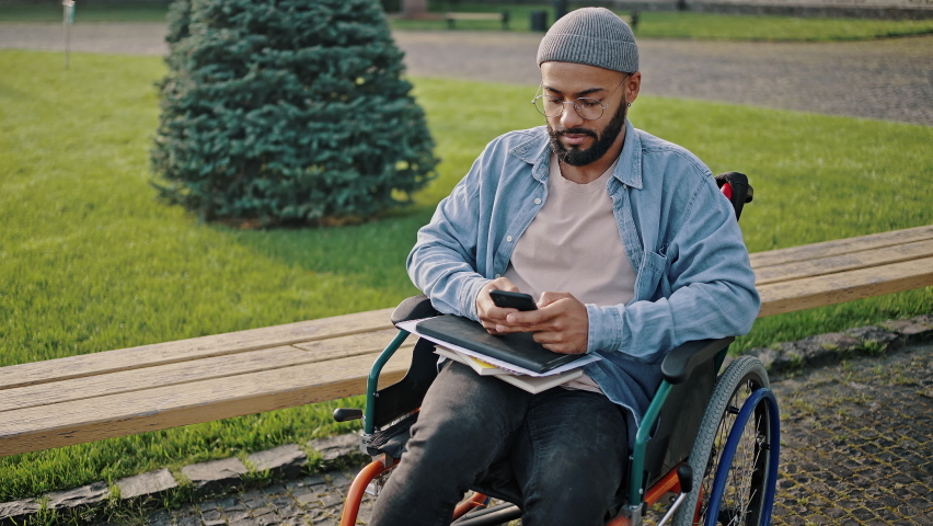 Disabled African American man reads message on smartphone from friend smiling. Young male student in denim jacket sits in wheelchair on university campus