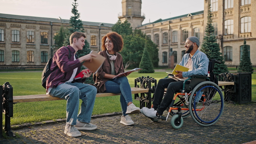 Man shows exercise book to disabled African American friend in wheelchair sitting on bench near black woman. Multinational friends prepare for classes slow motion | Shutterstock HD Video #1094464593