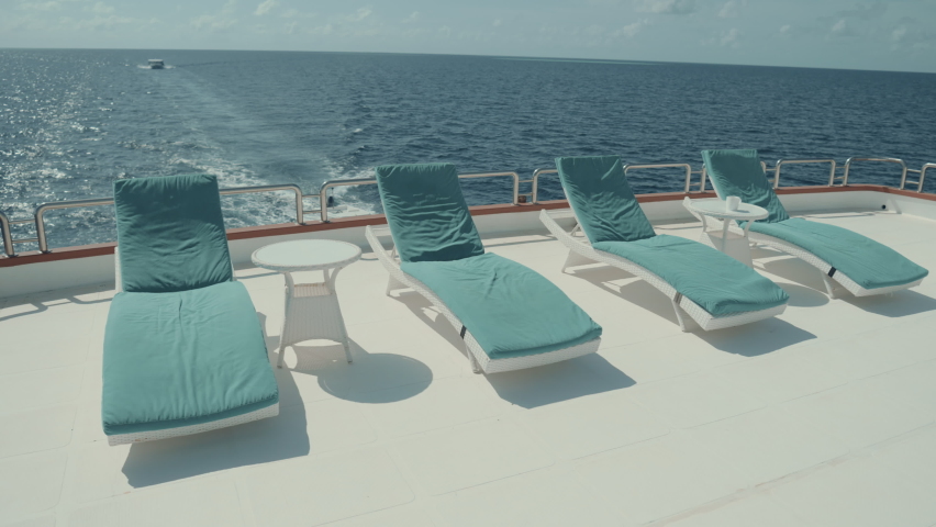 The row of daybeds on the upper deck of a moving luxurious diving safari yacht with small coffee tables in between and an ocean horizon in the background; several deck chairs on the top of a ship Royalty-Free Stock Footage #1094467669