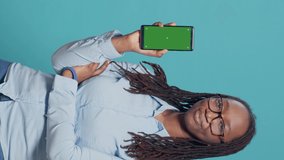 Vertical video: Female model holding smartphone with greenscreen display in studio, showing isolated mockup template with chromakey background on camera. Having mobile phone with blank copyspace.