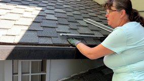 4K HD video of older caucasian woman wearing basic gardening gloves installing gutter guards under shingles of roof over rain gutters on house. View from behind person working.