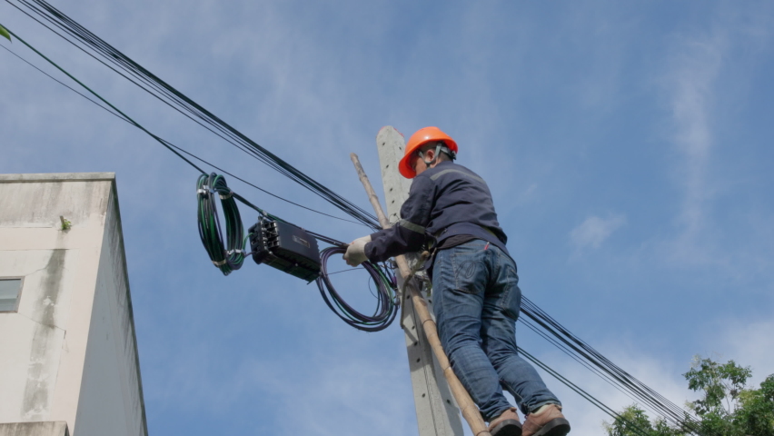 A telecoms worker is shown working from a utility pole ladder while wearing high visibility personal safety clothing, PPE, and a hard hat.

 | Shutterstock HD Video #1094473327