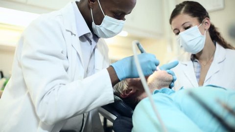 Dentist and dental nurse at work on a reclining patient. The dentist is using a dental probe, the nurse is busy with the suction tube. They are all three talking to each other.