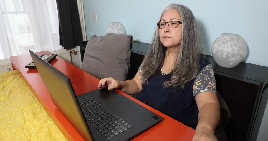 Senior adult woman sitting on bed using her laptop, conference speaking in front of webcam, facial expression of disagreement and happiness, long black gray hair, glasses. Concept of working at home Royalty-Free Stock Footage #1094478251