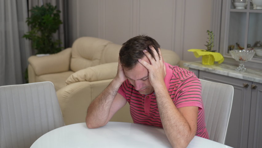 Depressed millennial man sitting at table in kitchen. Portrait of anxious handsome man having personal problems. Upset fired male employee sitting by table at kitchen and crying. | Shutterstock HD Video #1094478321