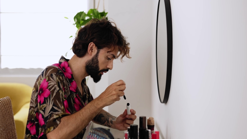 Young man applying eyelashes sitting at a dressing table. Transgender guy putting on makeup at home. LGTBI and diversity people concept. High quality 4k footage