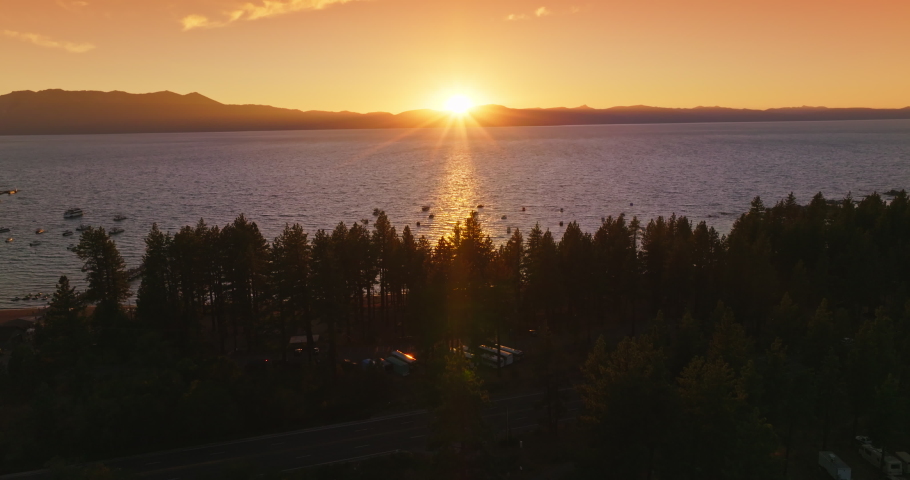 Dark silhouettes of pine trees on the waterfront of Lake Tahoe, California, USA. Numerous boats sailing on the water. Setting sun behind the mountains at backdrop.