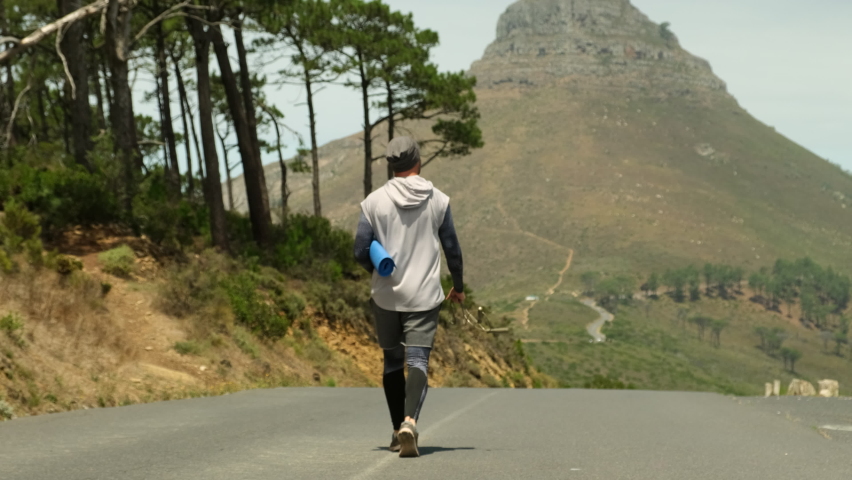 a sporty man in shorts and sports leggings walks along a road in a park near high mountains and holds a yoga mat and a jump rope in his hands. stylish sportsman man walking after jogging with yoga mat