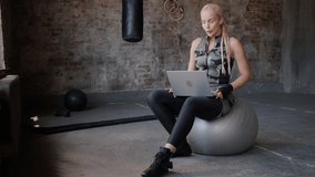 Professional coach uploads video of workout on site via laptop sitting on grey fitness ball in gym. Blonde woman uses modern device for work