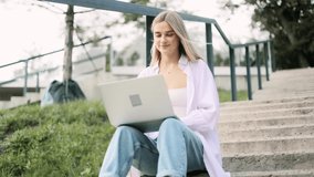 Attractive blonde woman sitting on stairs in the park and talking on laptop by video chat. Student learning online. Freelancer outside.