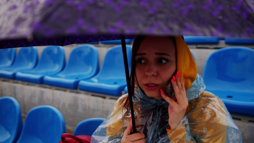 A beautiful woman in a hoodie and a plastic raincoat, talking on a mobile phone, sitting on the podium with an umbrella in her hand during the rain