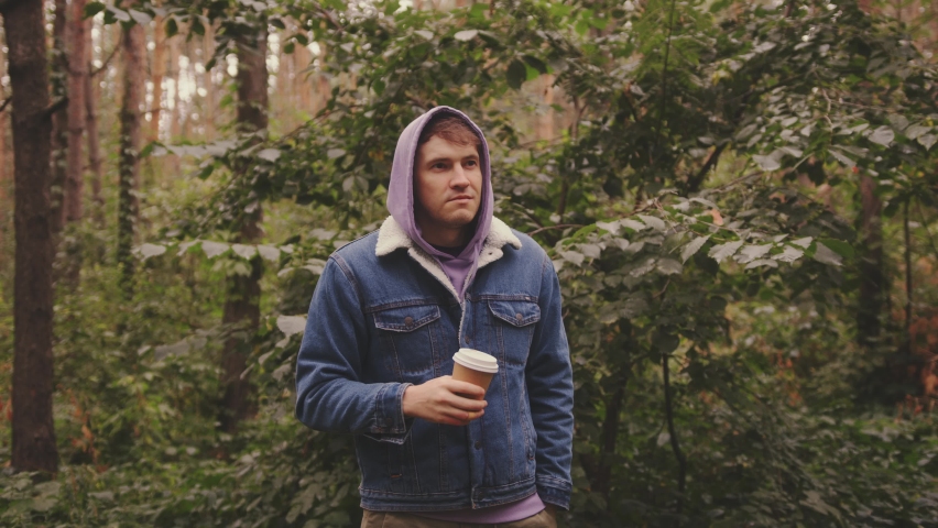 Young man in warm clothes drinking coffee from paper cup, standing in forest.