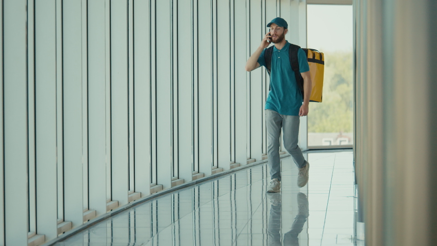 Young Food Delivery Person in Uniform and With a Thermal Backpack Walking in Modern Office Building and Using Smartphone to Check Order. Courier On the Way to Deliver Order to a Client.