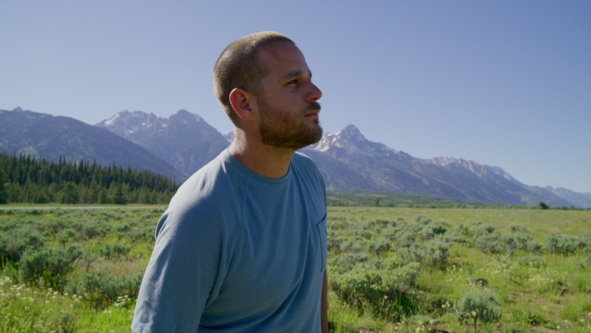 Male tourist in his 30s hiking along lush meadow with rising rocky range in the background. Solo traveler enjoys the serenity of iconic wilderness in the Grand Teton Park. High quality 4k footage
