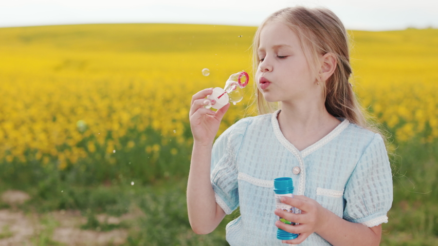 Adorable blond haired schoolgirl in stylish summer dress playing with soap bubbles on fresh air. Carefree little child with playful mood enjoying free time at countryside.