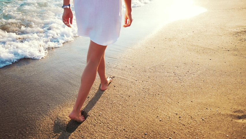 View back woman walks barefoot along shore beach and walks along sea. Sea water foam washes sand and feet of young beautiful tanned girl at sun rays sunset. Travel weekend on landscape outdoor nature. Royalty-Free Stock Footage #1094520227