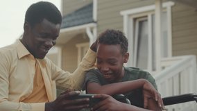 African American wheelchair boy playing games on smartphone together with his elder brother, sitting outside house in yard at summertime - Powered by Shutterstock - Get 15% off with code: PIKWIZARD15