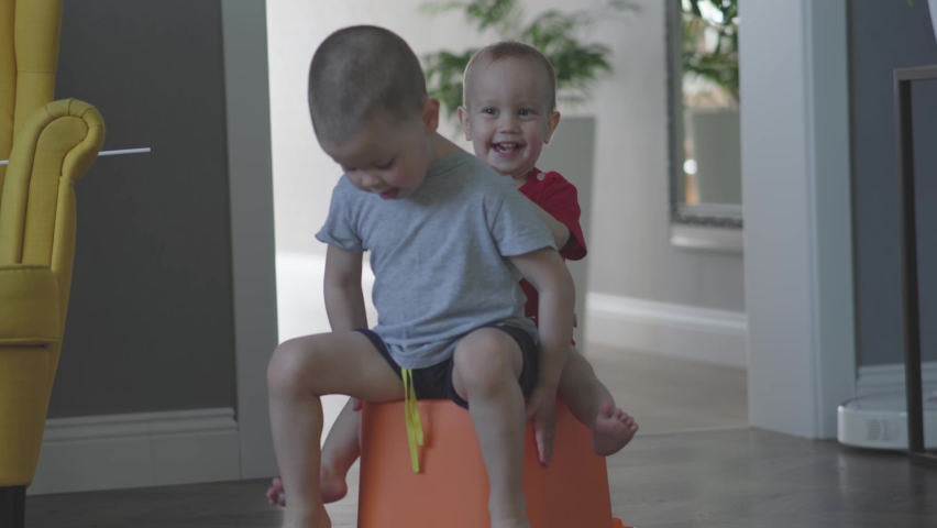little sibling brothers riding orange plastic box container, happy children playing together in living room. laughing smiling babies kids. elder brother driving rolling pushing, toddler having fun