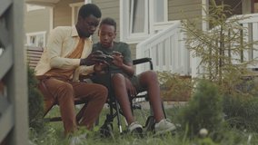 12 year old African American boy in wheelchair playing games on smartphone with his elder brother while spending time outdoors in yard outside house on warm sunny day - Powered by Shutterstock - Get 15% off with code: PIKWIZARD15