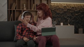 Mother putting on headphones to her autistic son sitting on the couch at home. Boy with autism using tablet computer with mom's help. - Powered by Shutterstock - Get 15% off with code: PIKWIZARD15