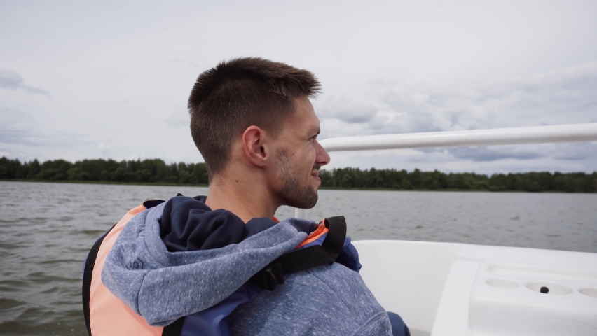 Middle aged caucasian man is sailing on pedal white boat on river on cloudy day and looking sideways. Brunette is talking, pedaling with feet and looking at shore with green trees.