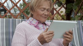 Elderly old caucasian woman outdoors surrounded by greenery is sitting in garden on bench swing. Communicates via video on tablet via Internet applications with family relatives and friends.