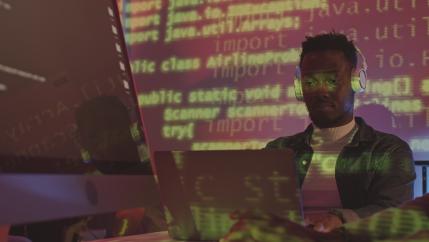 Waist up shot of young African American IT specialist in casualwear and headphones working on PC in office at night with green programming code lines projecting on his face | Shutterstock HD Video #1094532785