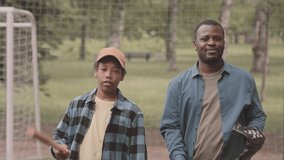 Medium portrait of African American dad with teenage son smiling at camera standing against net in park having baseball training outdoors in summer - Powered by Shutterstock - Get 15% off with code: PIKWIZARD15