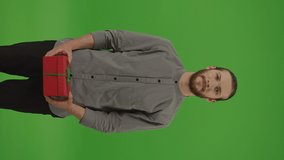 Vertical Video,Vertical View.Portrait of Young Motivated Bearded Man in Denim Shirt Holding Red Gift Box with Green Bow,Smiling and Presenting it on Green Screen,Chroma Key.Concept of Gift or Surprise