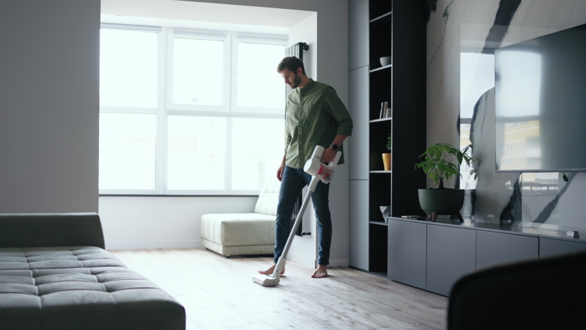 A bearded middle-aged man vacuums an apartment with a wireless hand-held vacuum cleaner against the background of a large window Royalty-Free Stock Footage #1094543533