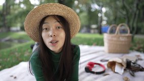 Portrait of charming Asian woman talking looking at camera pointing away smiling. Happy confident female blogger posing on summer picnic lying in park vlogging. Lifestyle and confidence concept
