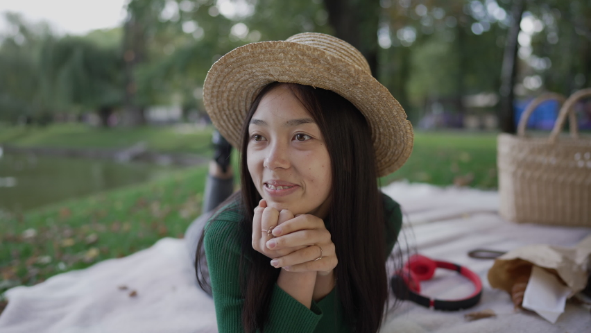 Joyful Asian young woman with teeth braces smiling looking away lying on blanket in park. Portrait of relaxed happy charming lady in straw hat enjoying weekend picnic outdoors on summer day