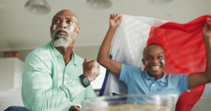 Video of african american father and son with flag of france watching match in tv. Family time, spending time together at home concept.