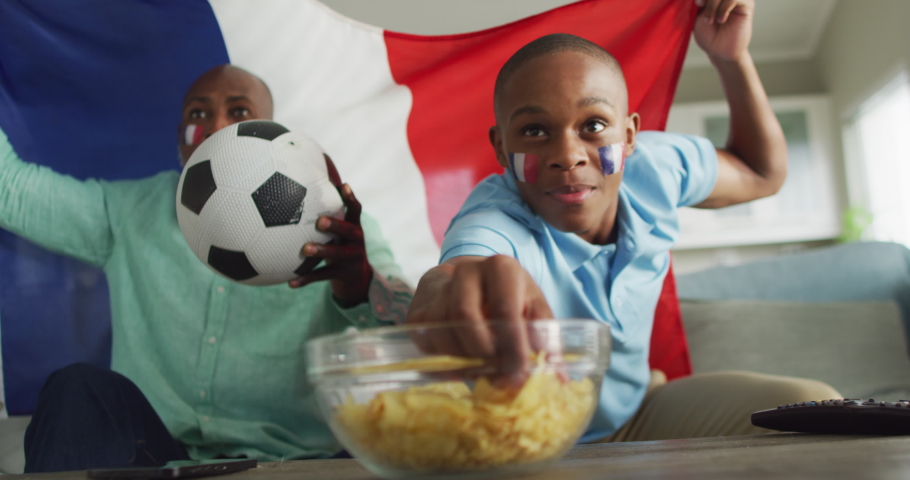 Video of happy african american father and son with soccer ball watching match. Family, spending quality time together at home.