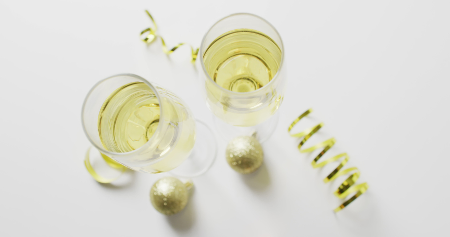 Overhead video of two full champgne glasses with gold streamers and baubles, on white background. New year