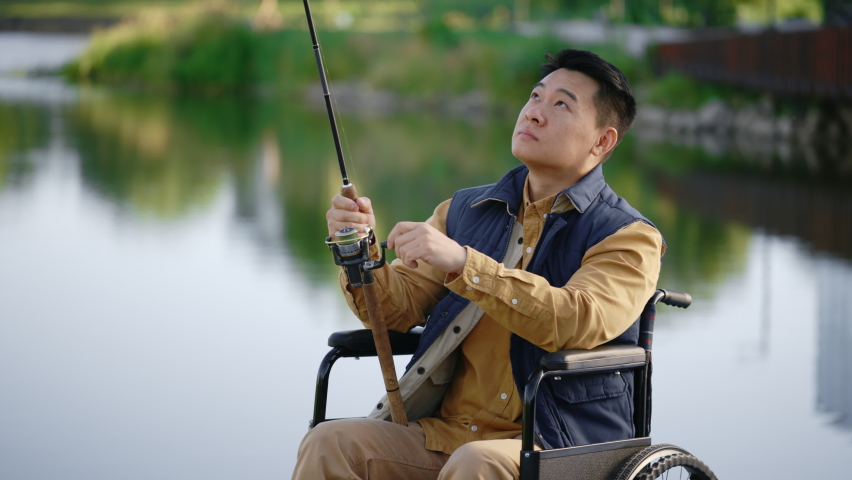 Portrait of disabled asian fisherman sitting on the wheelchair wearing special fishing clothes holding the fishing rod. Disabled people and fishing concept