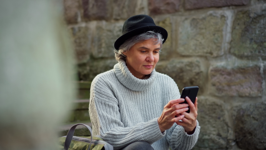 Portrait of mature woman sitting on stairs, using phone and smiling into camera. Camera raid, close up.