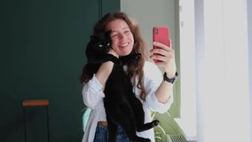 Smiling caucasian girl holding black pet cat using smartphone for video call, standing near window at home room. Sharing data on social media, using apps for virtual distant communication with friend