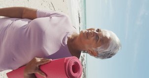 Vertical video of portrait of happy biracial woman with yoga mat on sunny beach. healthy, active retirement beach holiday.