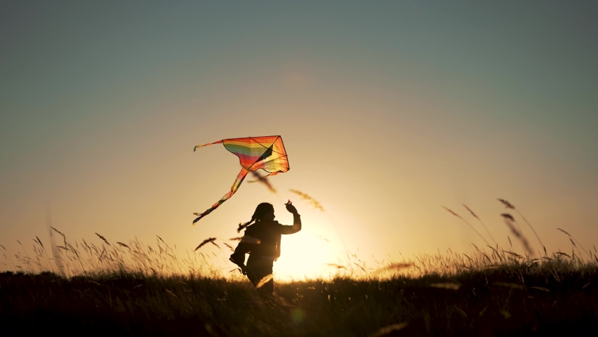 Happy child. Girl with flying kite in park. Child runs at sunset on grass in park. Silhouette of girl with flying kite. Child launches flying kite into sky in the wind. Silhouette of child in meadow Royalty-Free Stock Footage #1094574413